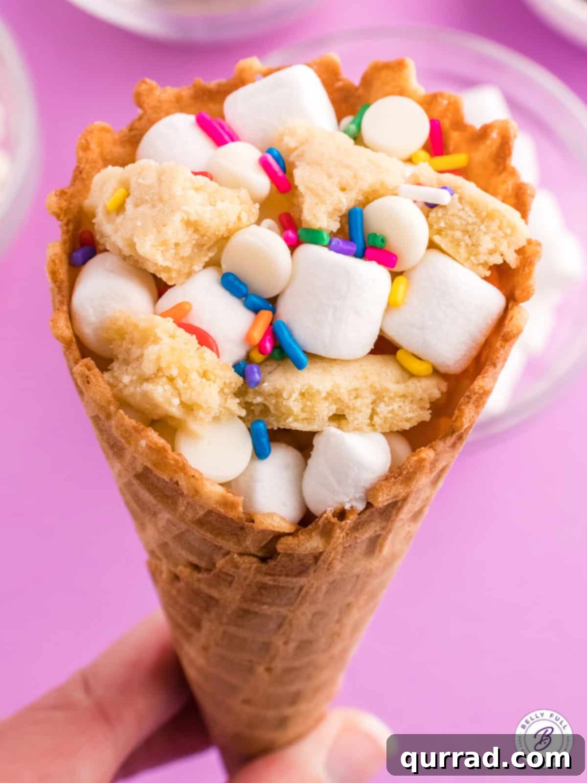 Gooey waffle cones stuffed with marshmallows, white chocolate chips, and sugar cookies – the perfect easy campfire treat! A hand holding a perfectly toasted sugar cookie campfire cone, with melted marshmallows and chocolate spilling slightly