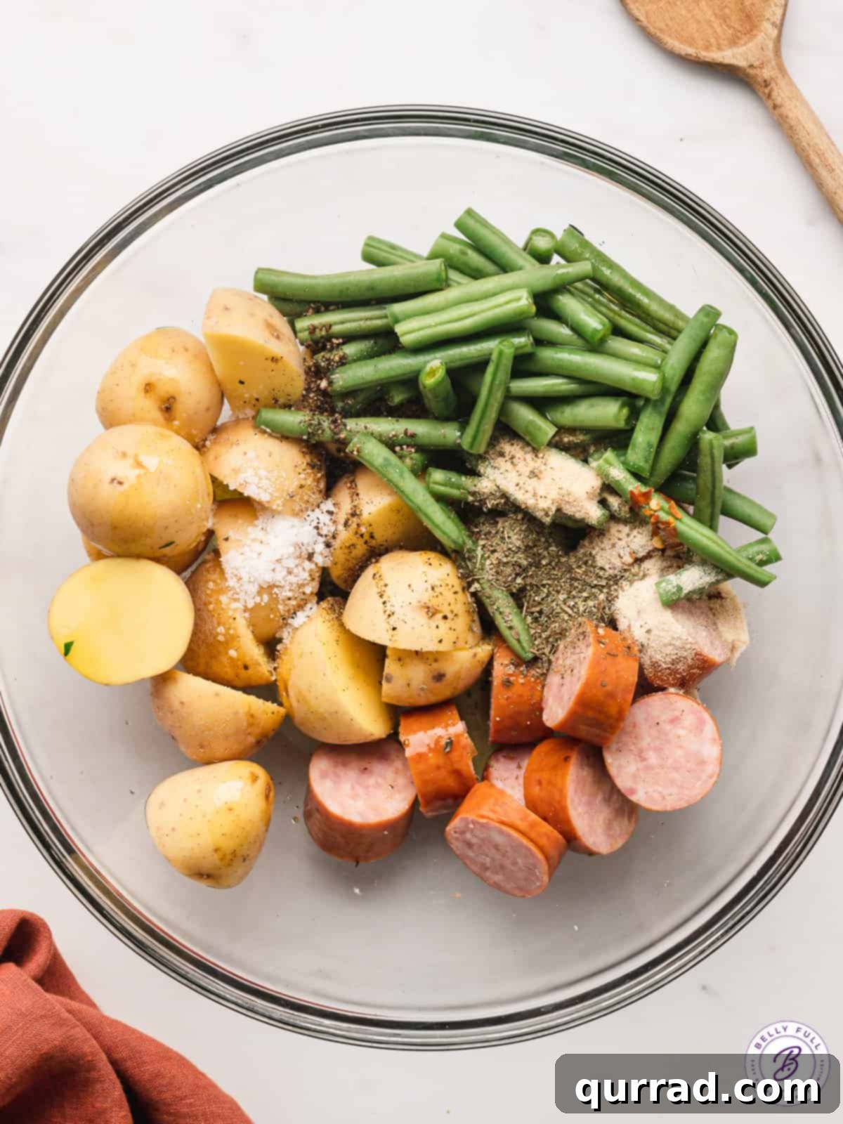 uncooked slices of kielbasa, potatoes, and green beans in bowl with seasonings