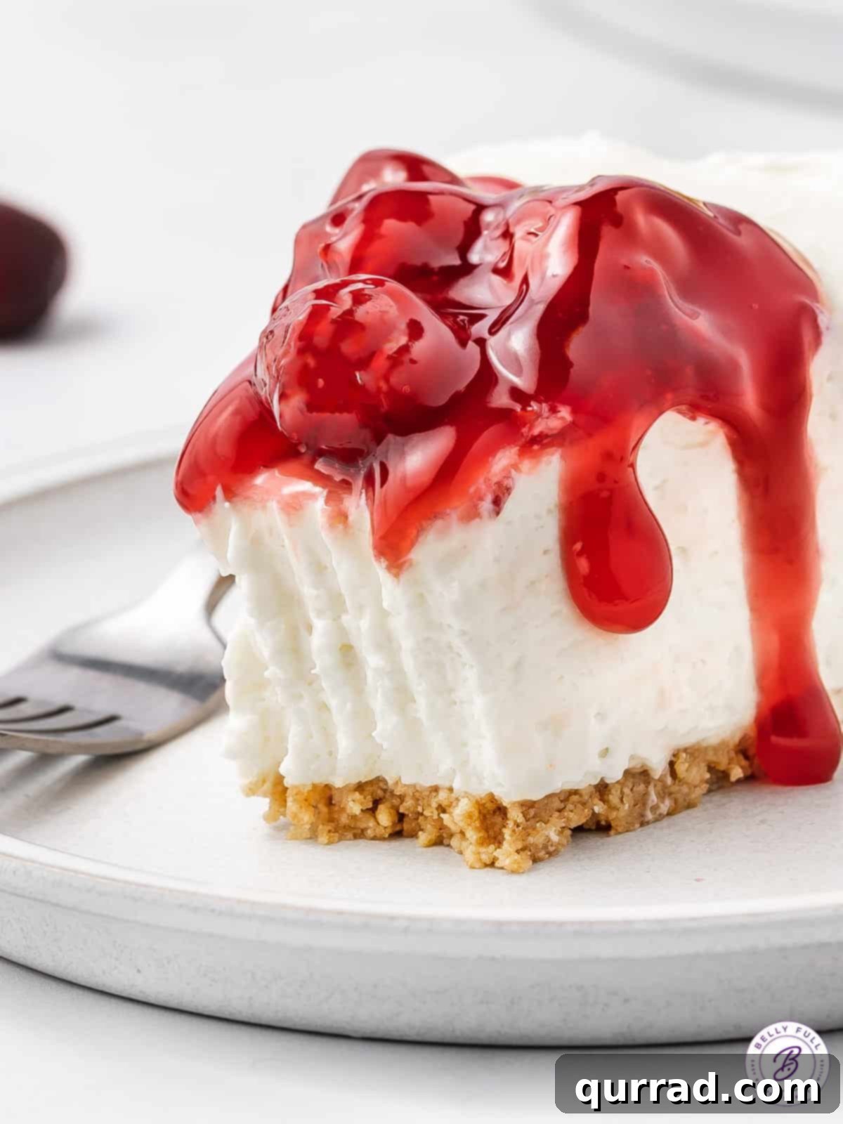With a tangy filling, graham cracker crust and cherry pie topping, this No Bake Cherry Cheesecake is the perfect balance of tart & sweet. A close-up shot of a single slice of easy cherry cheesecake on a white plate, with a small bite taken, revealing its creamy layers.