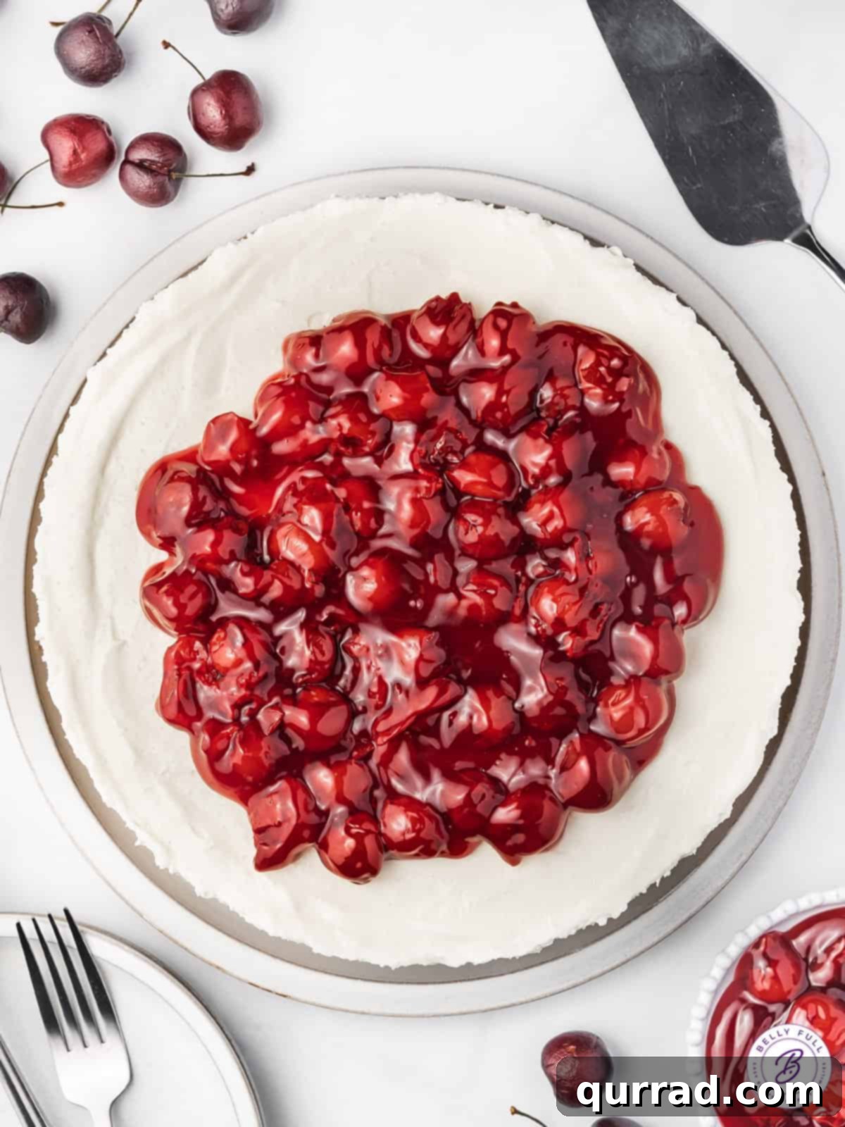 With a tangy filling, graham cracker crust and cherry pie topping, this No Bake Cherry Cheesecake is the perfect balance of tart & sweet. An overhead shot showcasing the vibrant red cherry pie filling evenly spread over the smooth, creamy surface of a no-bake cheesecake.