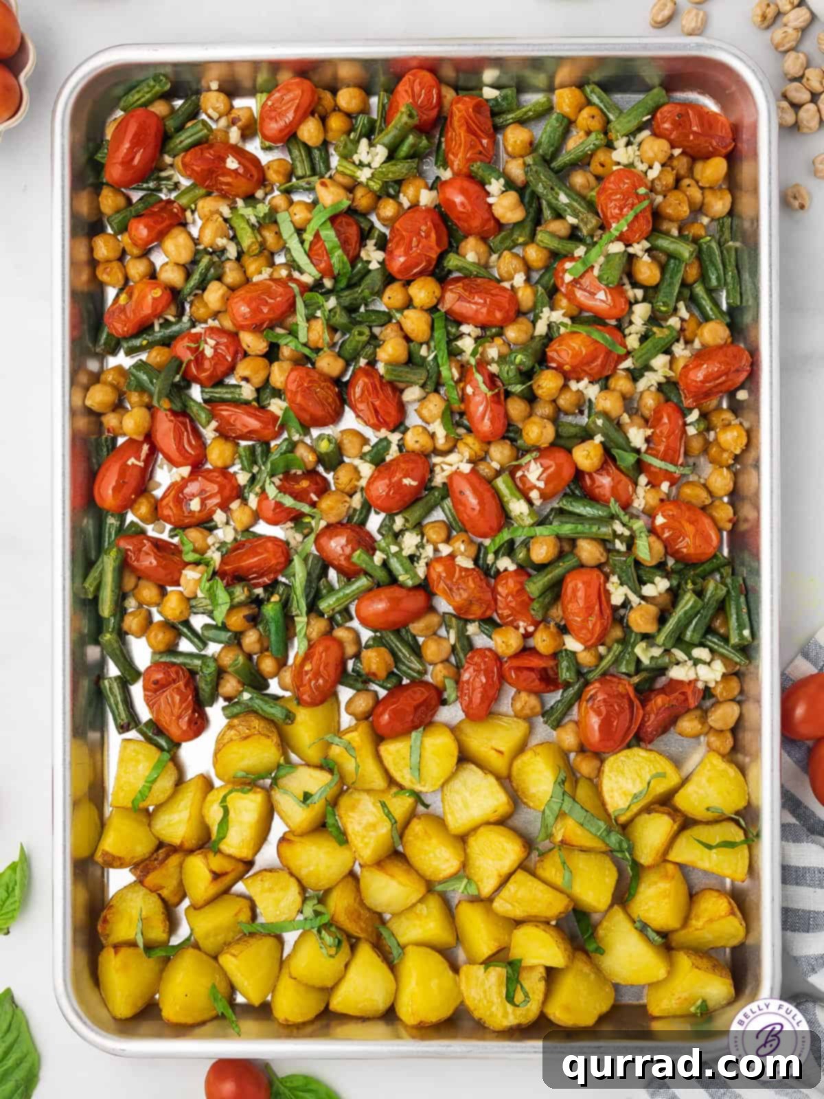 Close-up of chickpeas, cherry tomatoes, green beans, and quartered potatoes evenly spread on a baking sheet, ready for roasting.