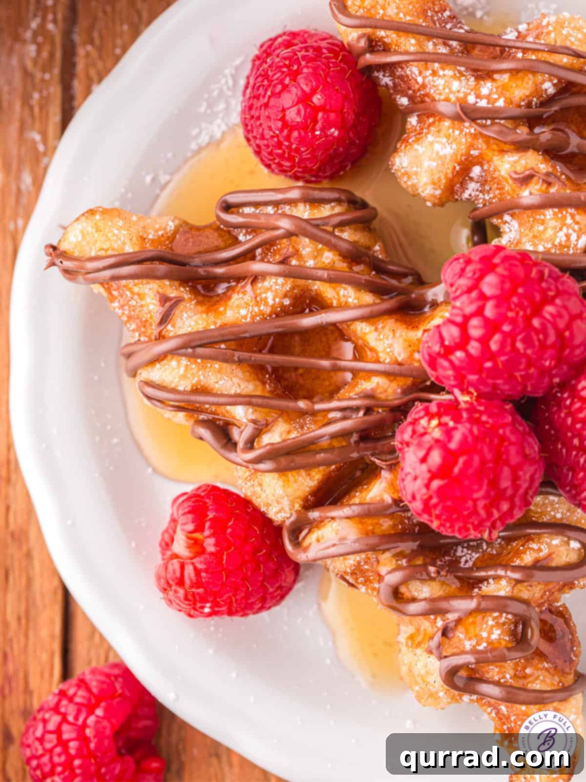 Freshly made croffles, drizzled with melted chocolate and garnished with vibrant red raspberries, presented invitingly on a plate.