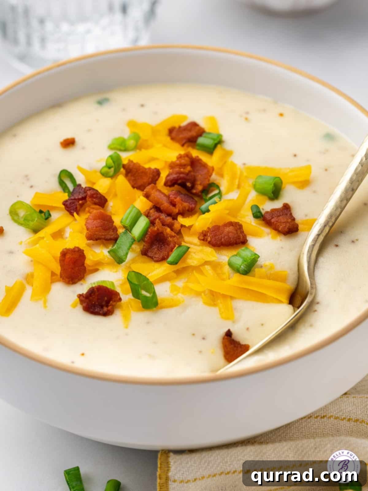 Copycat Panera Baked Potato Soup is rich, creamy, and full of incredible flavor, thanks to ingredients like cream cheese and bacon. Angled view of a bowl of creamy baked potato soup, generously topped with shredded cheddar cheese, crispy bacon bits, and fresh green scallions.
