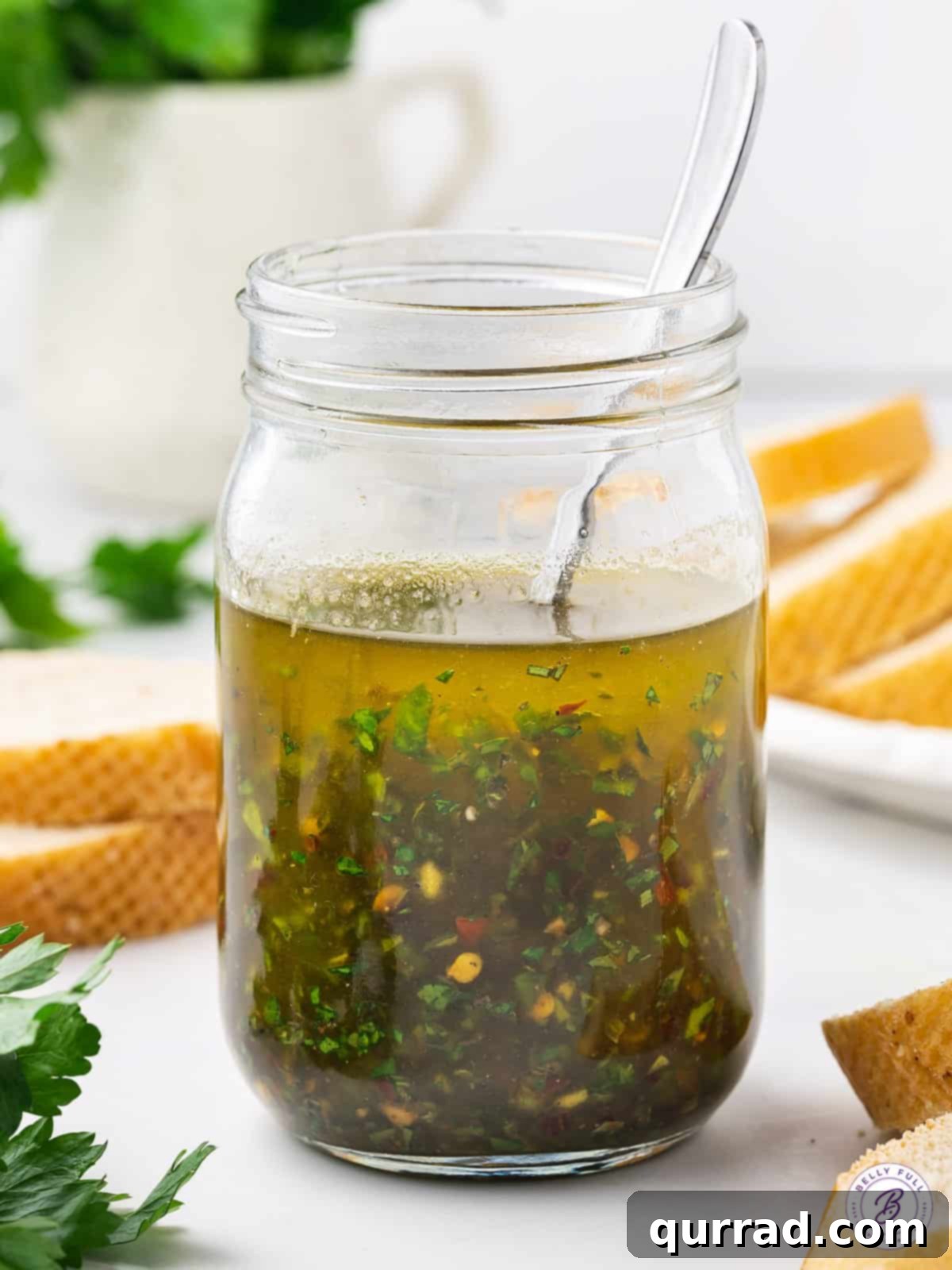 Close-up of high-quality extra virgin olive oil poured into a glass jar, surrounded by fresh herbs like parsley, thyme, and oregano, and small bowls of salt, black pepper, and red pepper flakes, highlighting the fresh ingredients for the dipping oil.