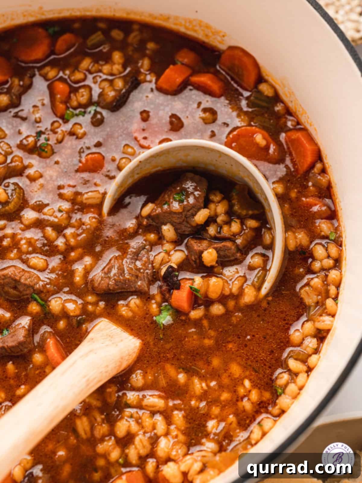 A ladle serving a generous portion of beef barley soup from a large pot.