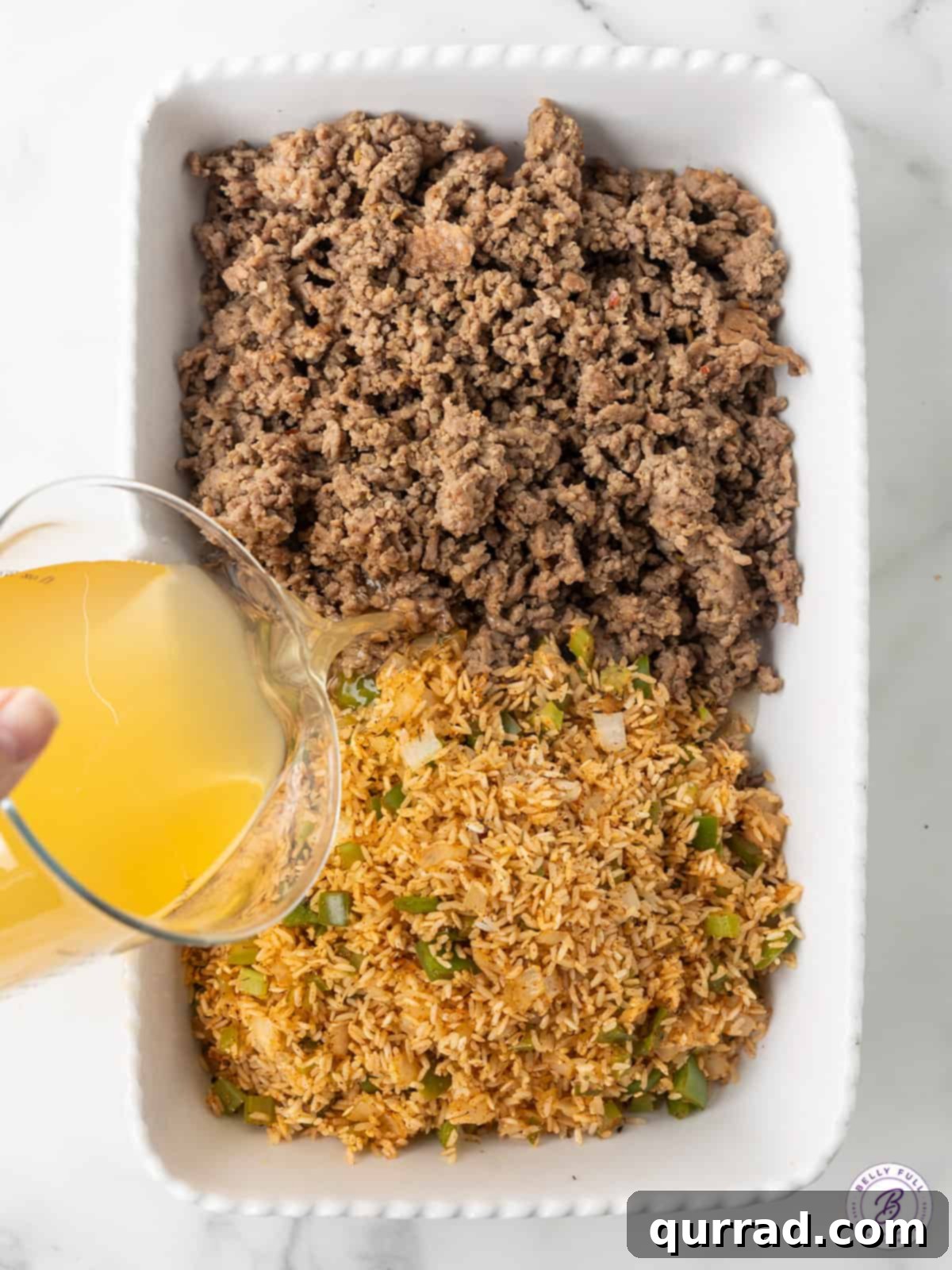 Chicken broth being poured over the cooked ground beef, rice, and vegetable mixture in a casserole dish, prior to baking.