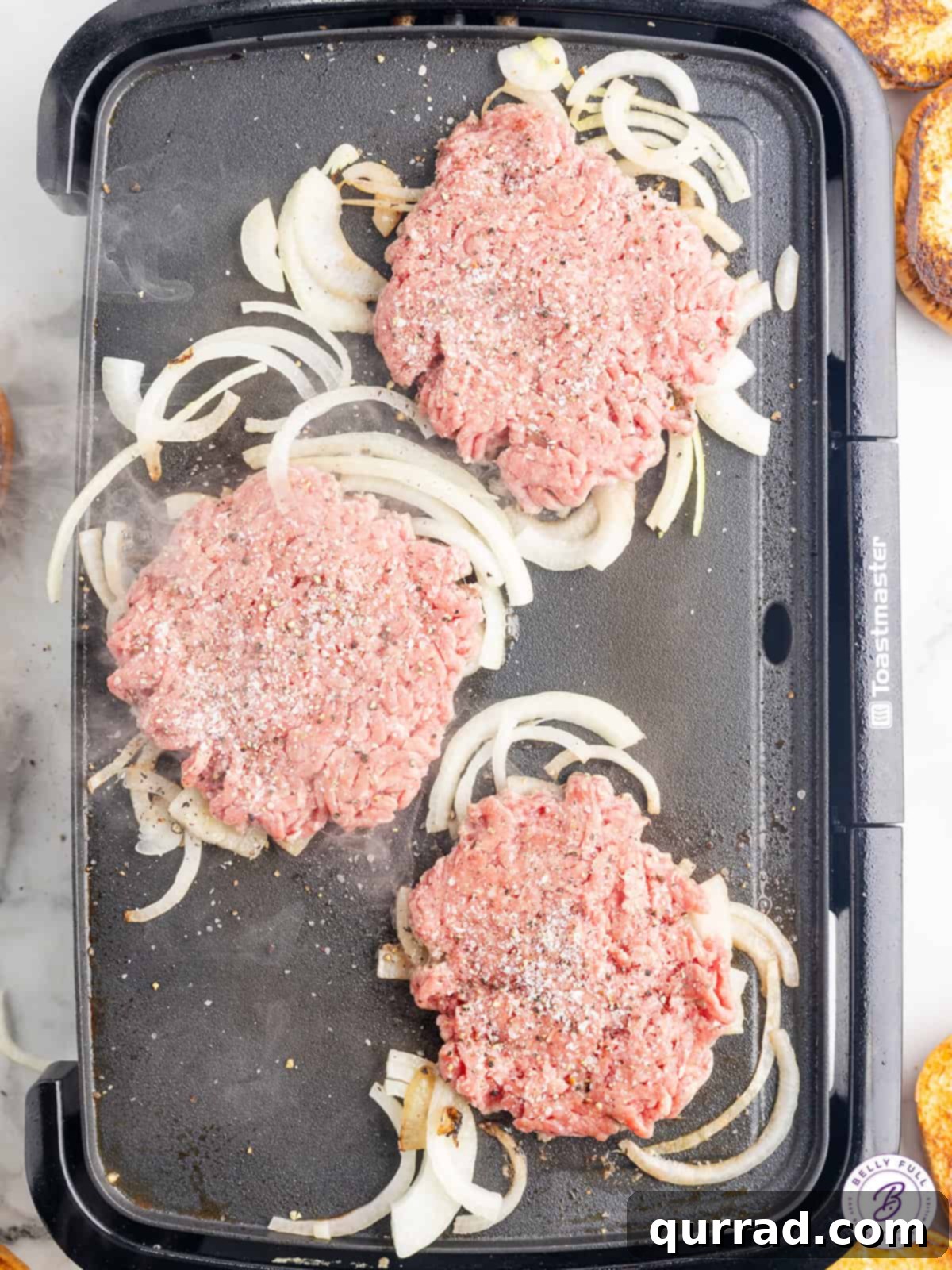 These Oklahoma Smash Burgers are burger patties smashed over thinly sliced onions, until the edges are crisp and the center remains juicy. Close up shot of burger patties being smashed onto thinly sliced onions on a hot griddle, showing the process of creating Oklahoma smash burgers.