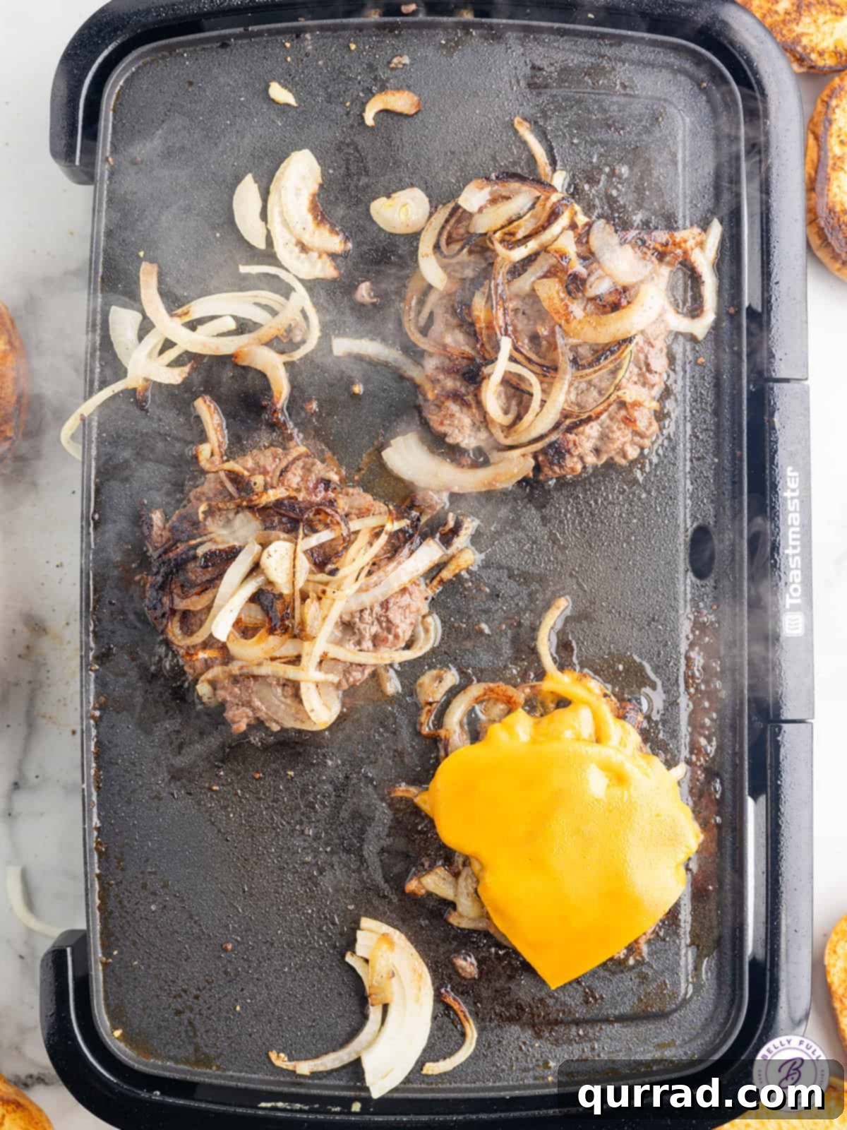 These Oklahoma Smash Burgers are burger patties smashed over thinly sliced onions, until the edges are crisp and the center remains juicy. Oklahoma smash burgers cooking on a griddle, with one patty topped with a slice of melting American cheese.