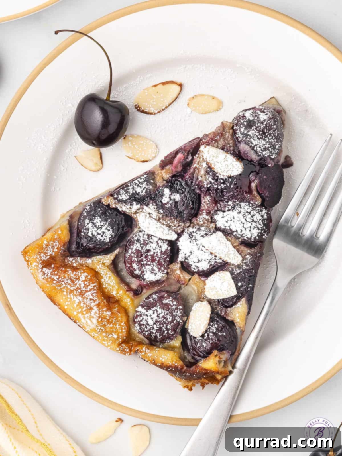 This Cherry Clafoutis recipe features a rich custard-like filling dotted with fresh tart cherries. Easy yet elegant! A slice of cherry clafoutis on a white plate
