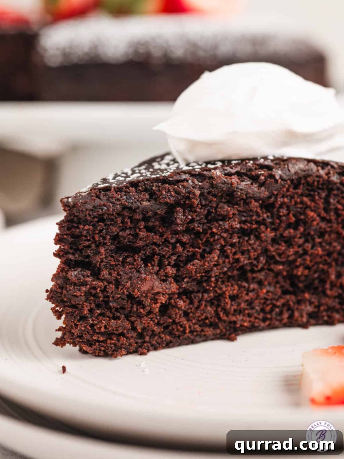 Enjoy a super moist Vegan Chocolate Cake, made with cocoa powder, chopped chocolate, and almond milk, beautifully paired with whipped cream. A slice of vegan chocolate cake artfully topped with fluffy whipped cream and fresh berries on a delicate plate.
