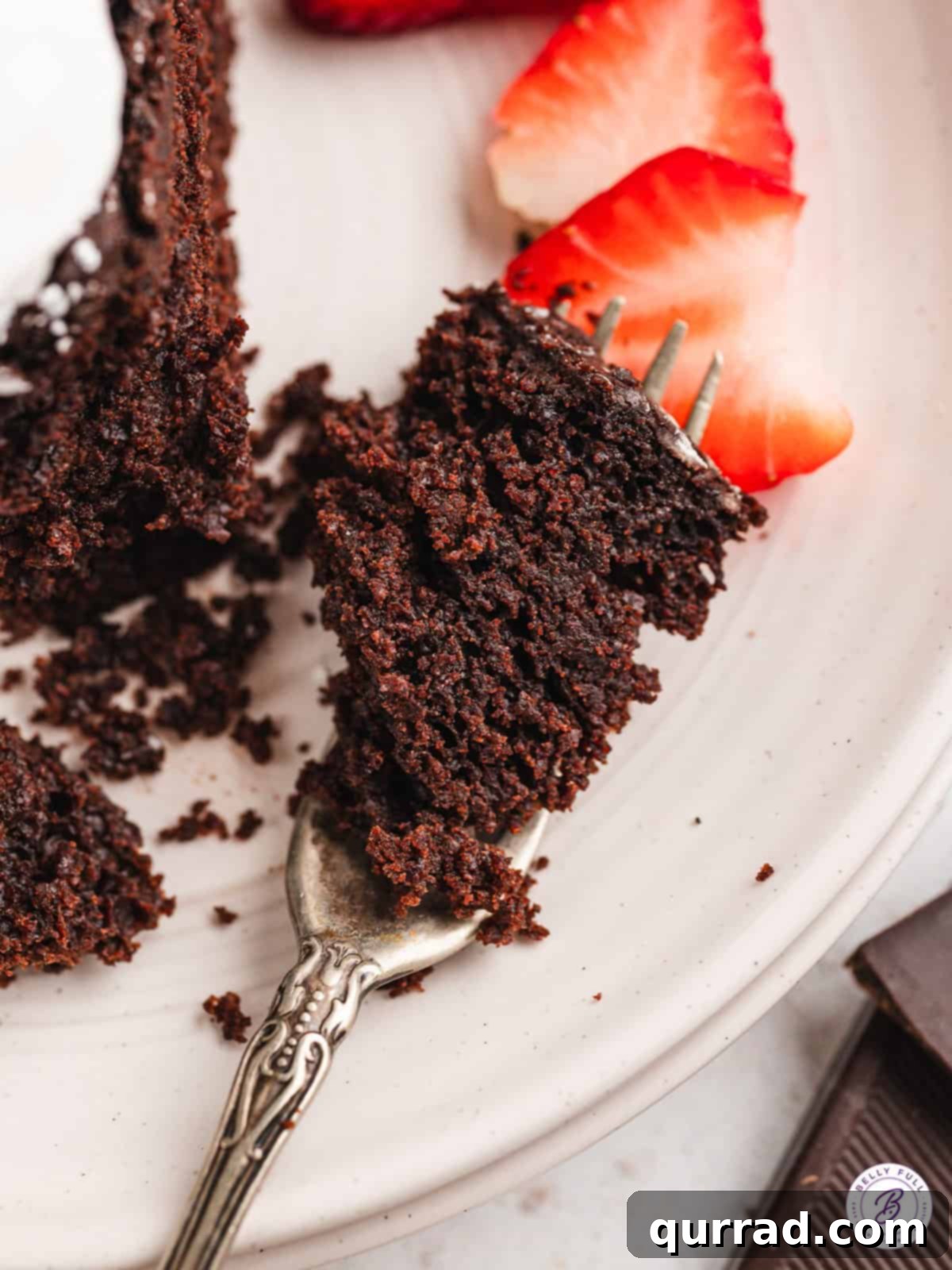 Savor every bite of this super moist Vegan Chocolate Cake, crafted with cocoa powder, chopped chocolate, and almond milk for a truly rich flavor. A tantalizing close-up of a bite of vegan chocolate cake on a fork, resting on a plate, highlighting its moist and rich interior.
