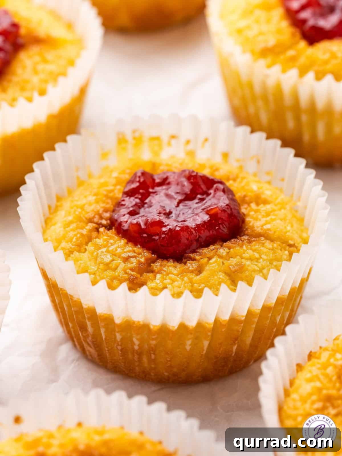 A cross between a creamy custard tart and an eggy macaroon, these Mini Coconut Cakes are a popular Portuguese dessert. A golden-brown Portuguese Mini Coconut Cake topped with a vibrant red raspberry jam, showcasing its creamy texture.