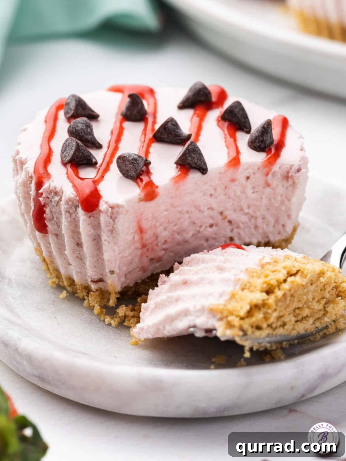 These No Bake Mini Strawberry Cheesecakes are sweet, tart, and irresistible. The filling is smooth, creamy and light, thanks to the Cool Whip. A miniature strawberry cheesecake with a bite on the fork next to it, showing its creamy texture and fresh strawberry topping.