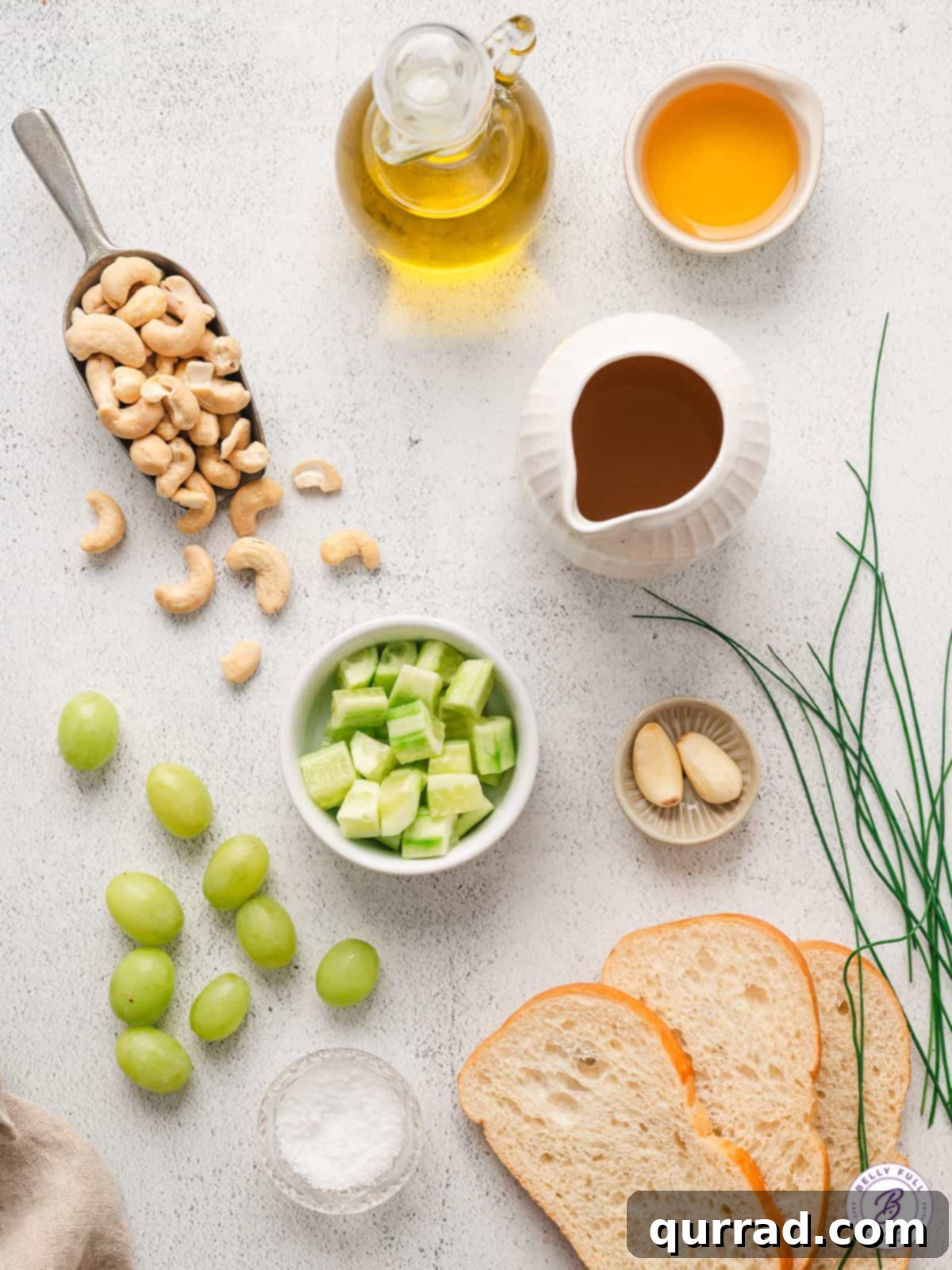 An assortment of fresh ingredients for cold cucumber soup laid out on a rustic surface, including whole cucumbers, green grapes, garlic cloves, bread slices, and a bottle of olive oil.