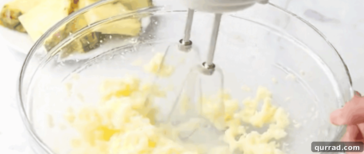 A close-up shot of a hand-held electric mixer vigorously combining softened butter and granulated sugar in a clear glass mixing bowl, creating a light and fluffy base for the pineapple bread batter.