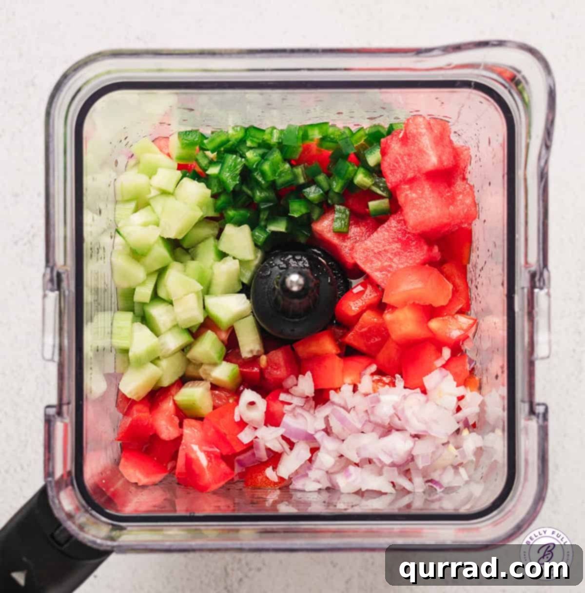 Overhead view of various fresh ingredients for watermelon gazpacho, including watermelon cubes, sliced cucumbers, tomatoes, bell peppers, and herbs, arranged around a blender.