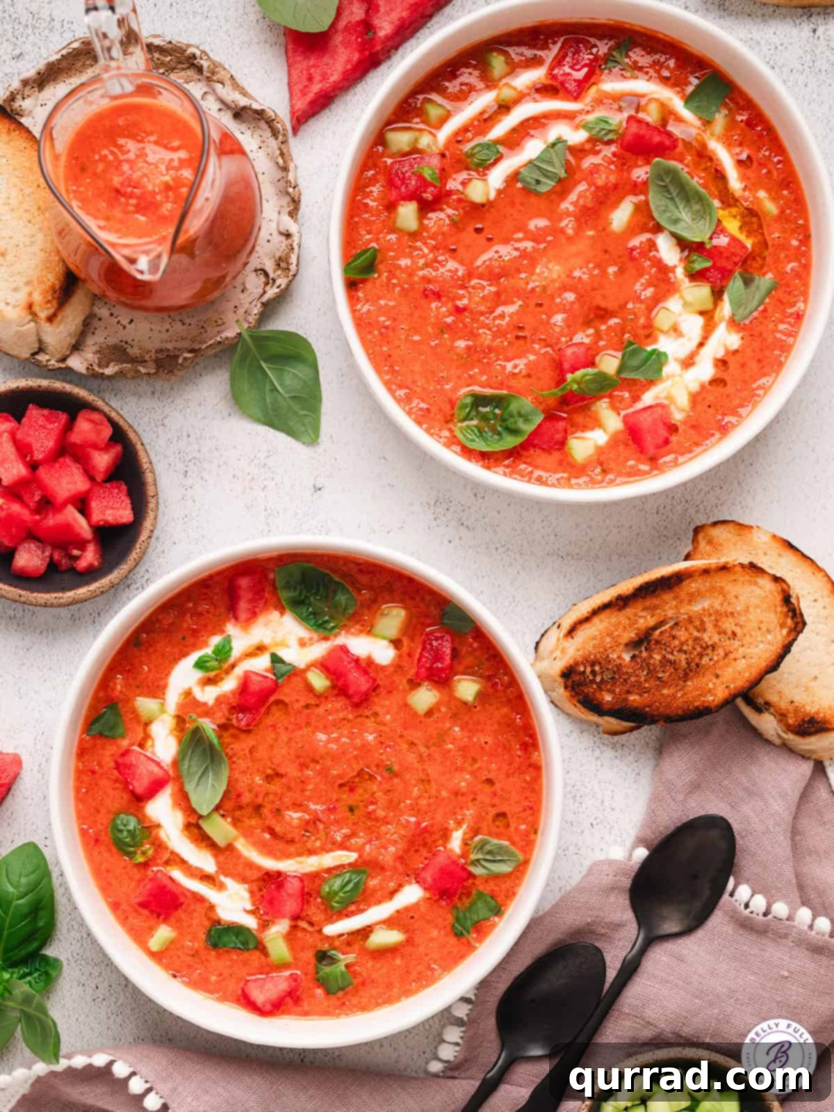 Two bowls of watermelon gazpacho, garnished with fresh herbs and diced cucumber and watermelon, on a rustic wooden surface.