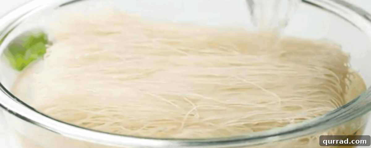 A hand pouring hot water over dry Thai Vermicelli rice noodles in a clear glass bowl, preparing them for the Pancit dish.