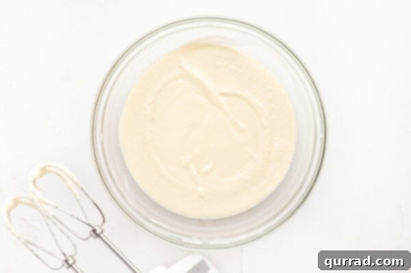 A creamy mixture of cream cheese, heavy cream, powdered sugar, and butter being whipped in a glass mixing bowl.