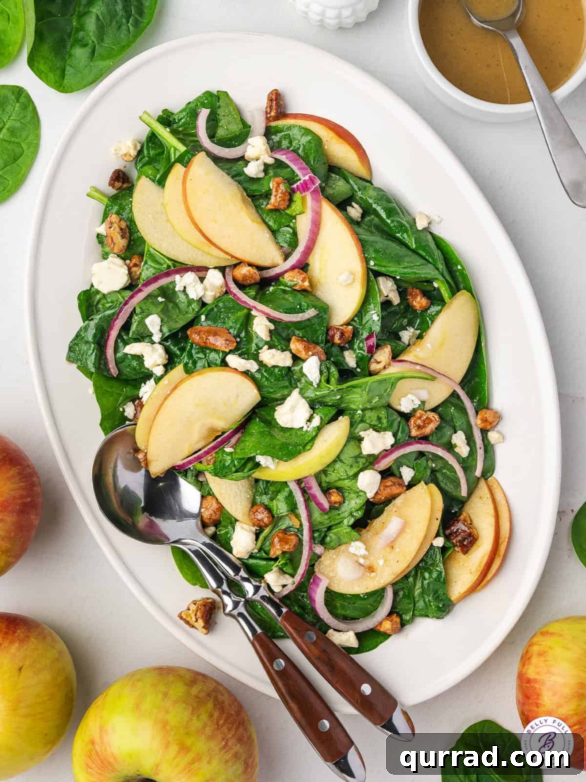 Overhead view of a vibrant spinach apple salad on a white platter, garnished with goat cheese and candied pecans.