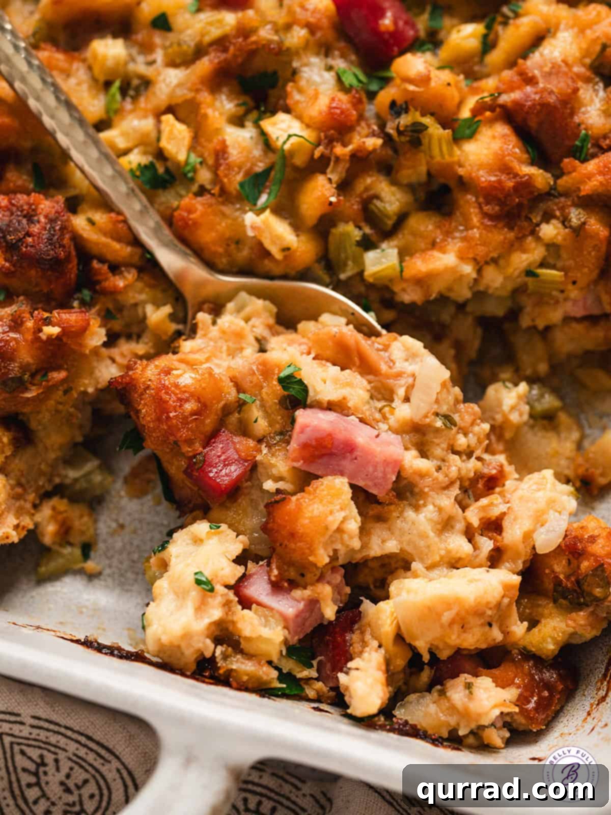 Close up of a spoon in a dish of potato bread stuffing, showcasing ham, cheese, and apple pieces