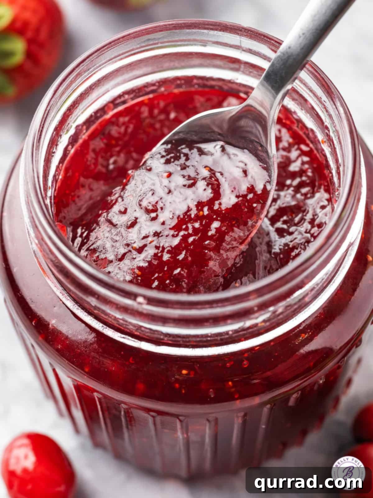 Made with strawberries, cranberries, and orange, this Christmas Jam is sweet, tart, and festive. Enjoy on biscuits or give as a gift. A spoon dipping into an open jar of festive Christmas jam, showcasing its rich texture and color.