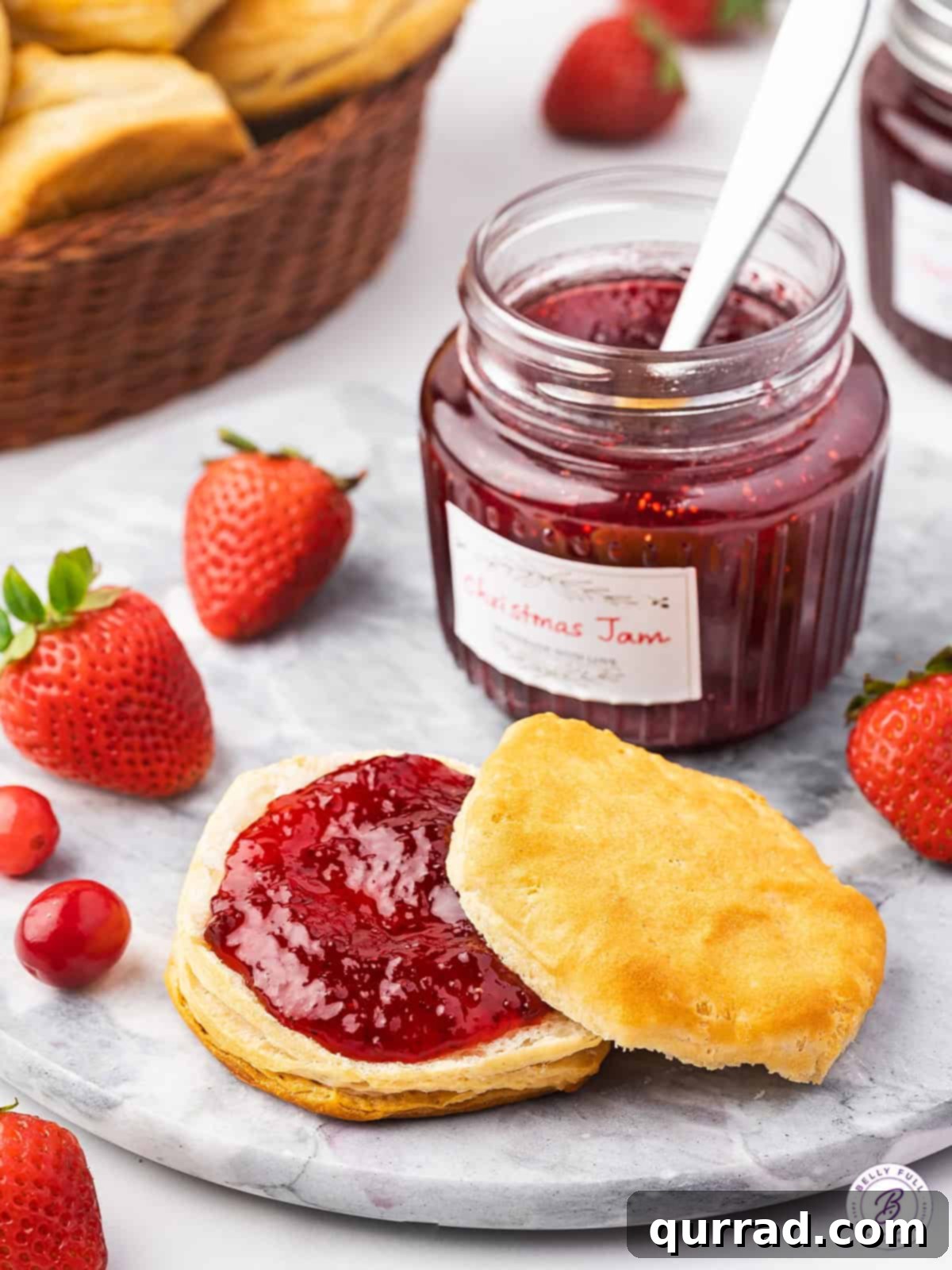 Made with strawberries, cranberries, and orange, this Christmas Jam is sweet, tart, and festive. Enjoy on biscuits or give as a gift. A warm biscuit generously topped with Christmas jam, with a jar of jam visible in the background.
