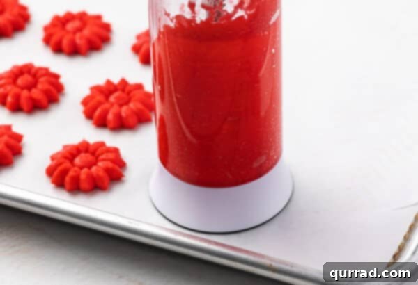 A spritz cookie press extruding red flower-shaped cookies onto a parchment-lined baking sheet.