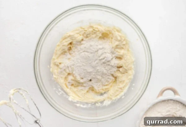 Flour being added gradually to the wet ingredients for spritz cookies in a glass mixing bowl.