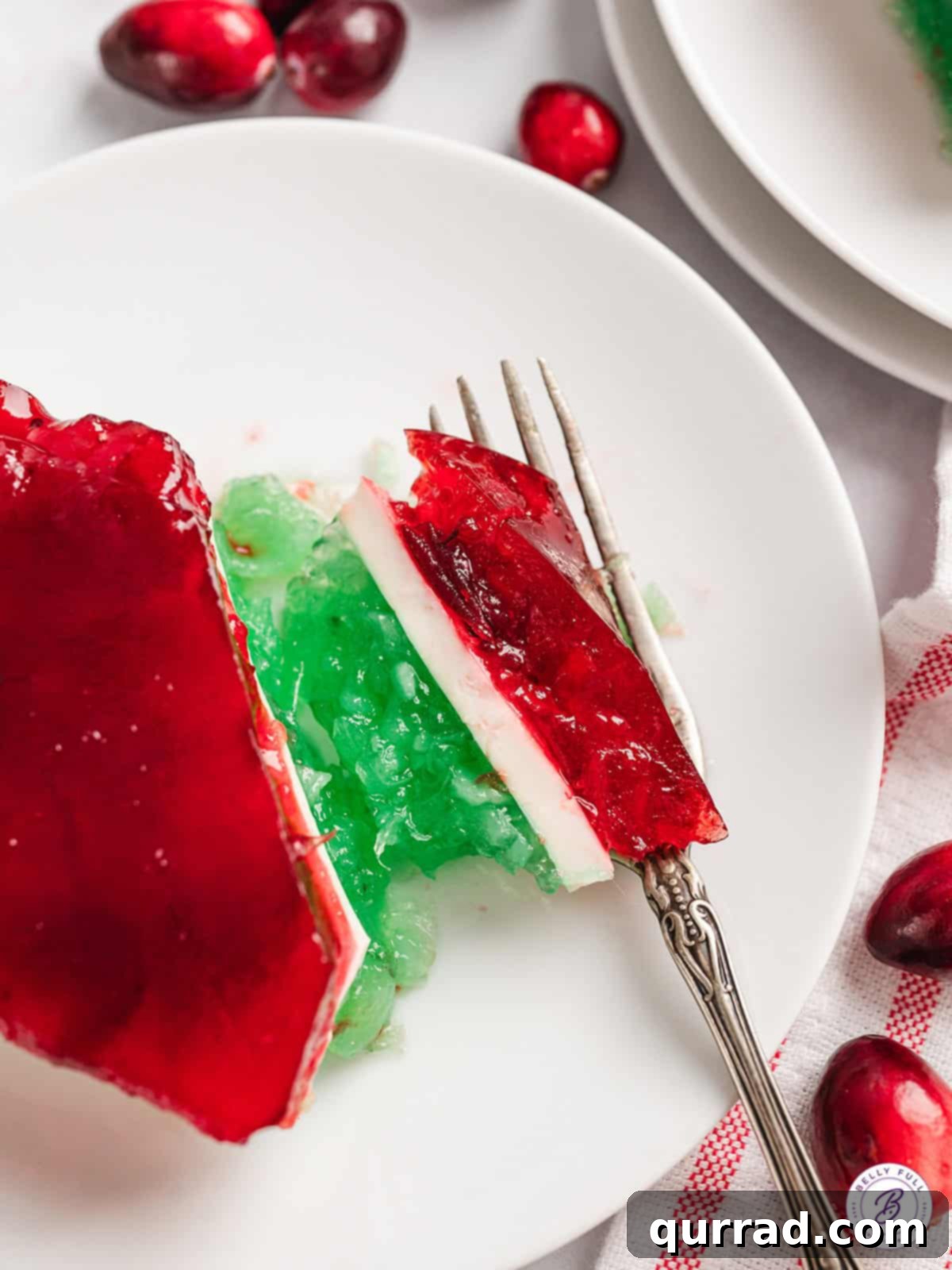 Close-up of Layered Christmas Jello Slice A perfectly sliced piece of layered Christmas jello on a white plate, with a fork resting beside it, highlighting the distinct green, white, and red layers.