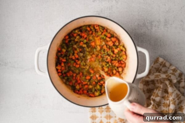 Pouring chicken broth into a stock pot containing sautéed vegetables and spices, creating the liquid base for the soup.
