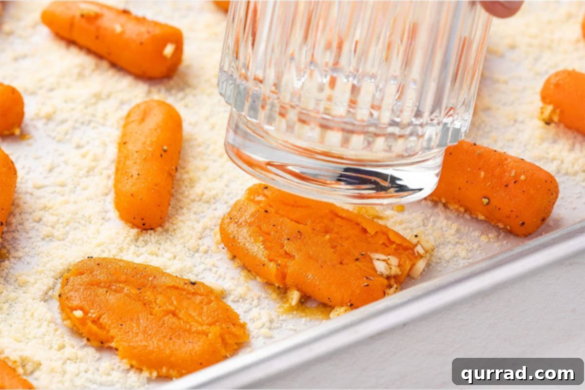 A glass pressing down on pre-cooked carrots on a parchment-lined baking sheet, demonstrating the smashing process