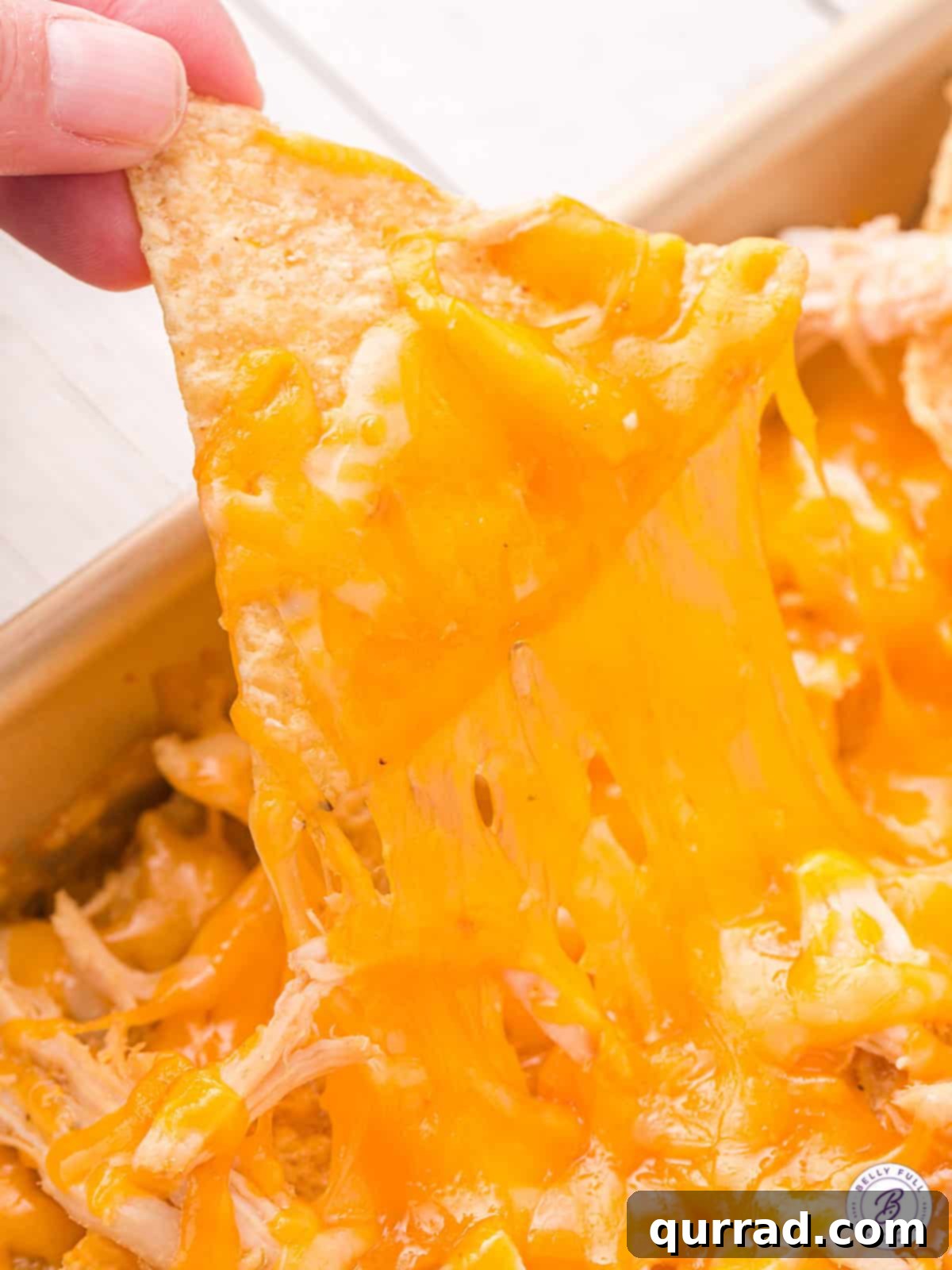 Layers of chips, cheese, and shredded chicken are baked and topped with all your favorite toppings in this Chicken Nachos recipe. A close-up shot of a hand pulling a cheesy chicken nacho away from the baking pan, highlighting the melted cheese pull.