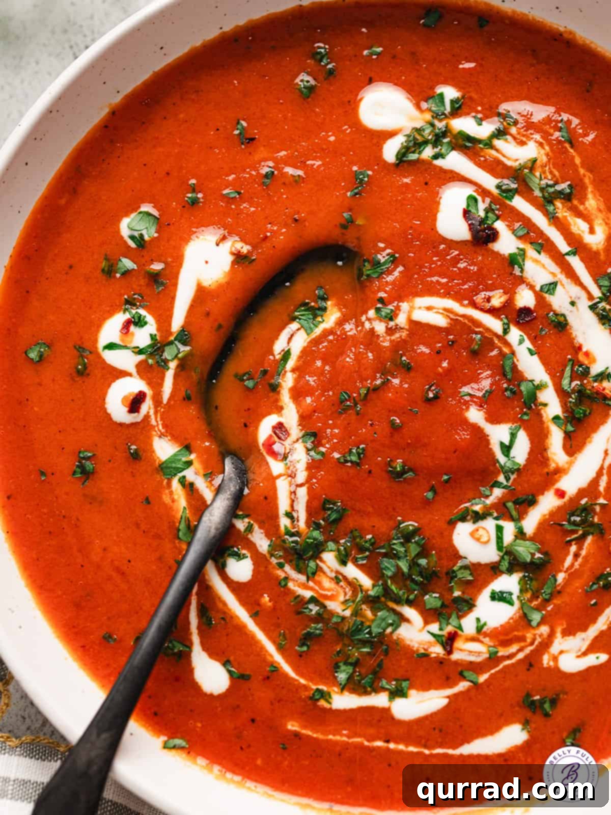 Overhead close up of a bowl of roasted red pepper soup, topped with cream and fresh parsley, with a spoon.