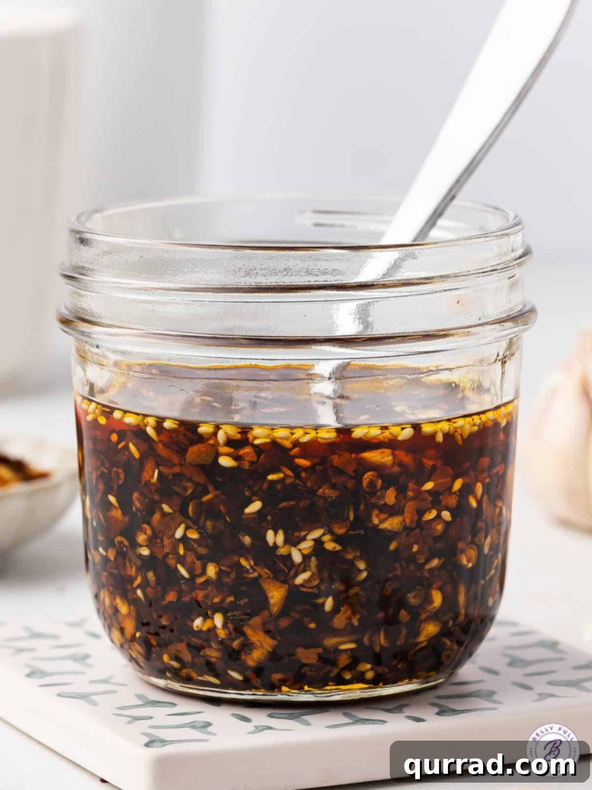 A spoon scooping homemade chili crisp from a clear glass jar, showing its rich texture.