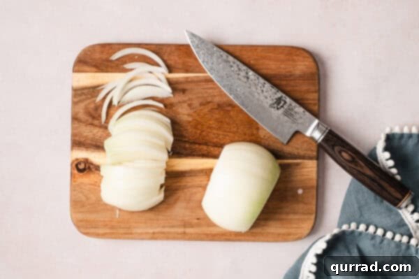 An onion on a cutting board, half of it thinly sliced for caramelization.
