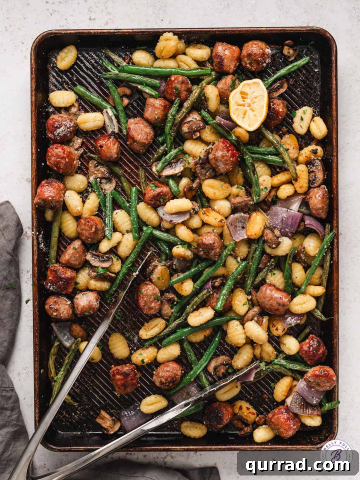 This Sheet Pan Gnocchi and Sausage is loaded with veggies and perfectly seasoned for an easy weeknight dinner the entire family will love. Tongs on a sheet pan of sausage and gnocchi, showing a delicious mix of ingredients