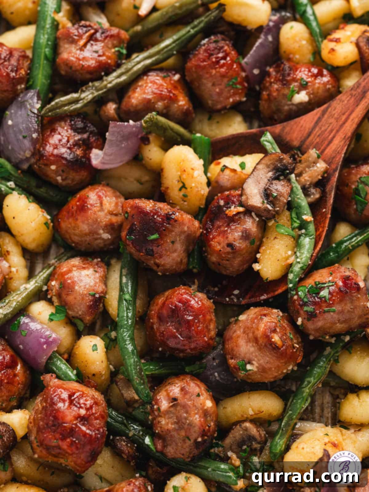 This Sheet Pan Gnocchi and Sausage is loaded with veggies and perfectly seasoned for an easy weeknight dinner the entire family will love. Close up of a wooden spoon stirring sheet pan gnocchi, showcasing crispy gnocchi and vibrant vegetables