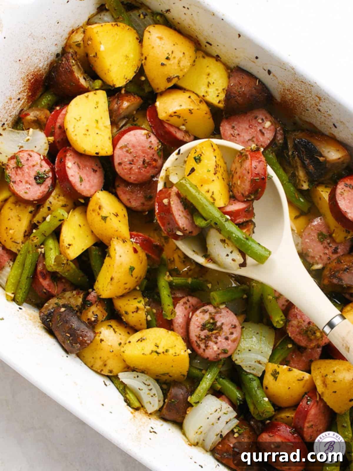 Close-up overhead view of the baked kielbasa and mixed vegetables, including mushrooms, onions, and green beans, glistening in the casserole dish with a serving spoon tucked in.