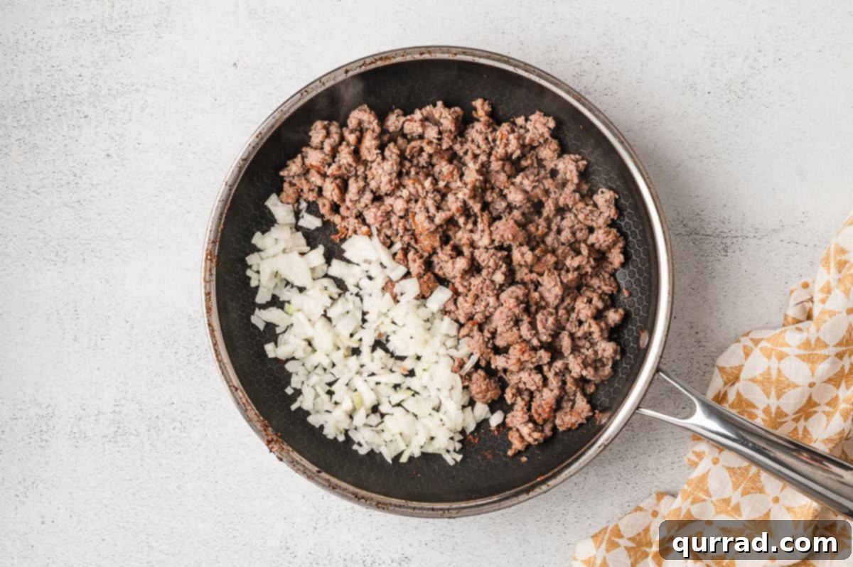 Crumbled sausage and diced onions cooking in a frying pan until softened and lightly browned.