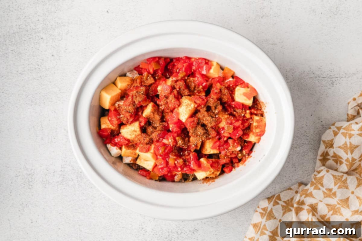 An undrained can of Rotel tomatoes and green chilies being poured over cubed Velveeta and cream cheese in a crockpot.