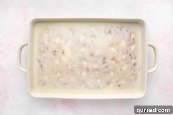 The creamy white filling being poured and drizzled over the baked cake, with visible liquid seeping into the poked holes.