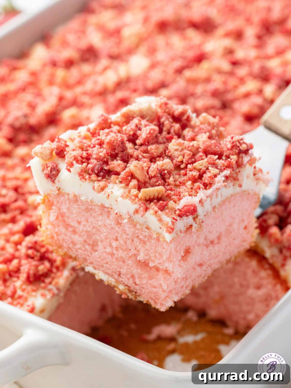 Strawberry cake is topped with homemade vanilla icing and a freeze-dried strawberry topping in this easy Strawberry Crunch Cake recipe. Lifting a slice of strawberry crunch cake from the pan