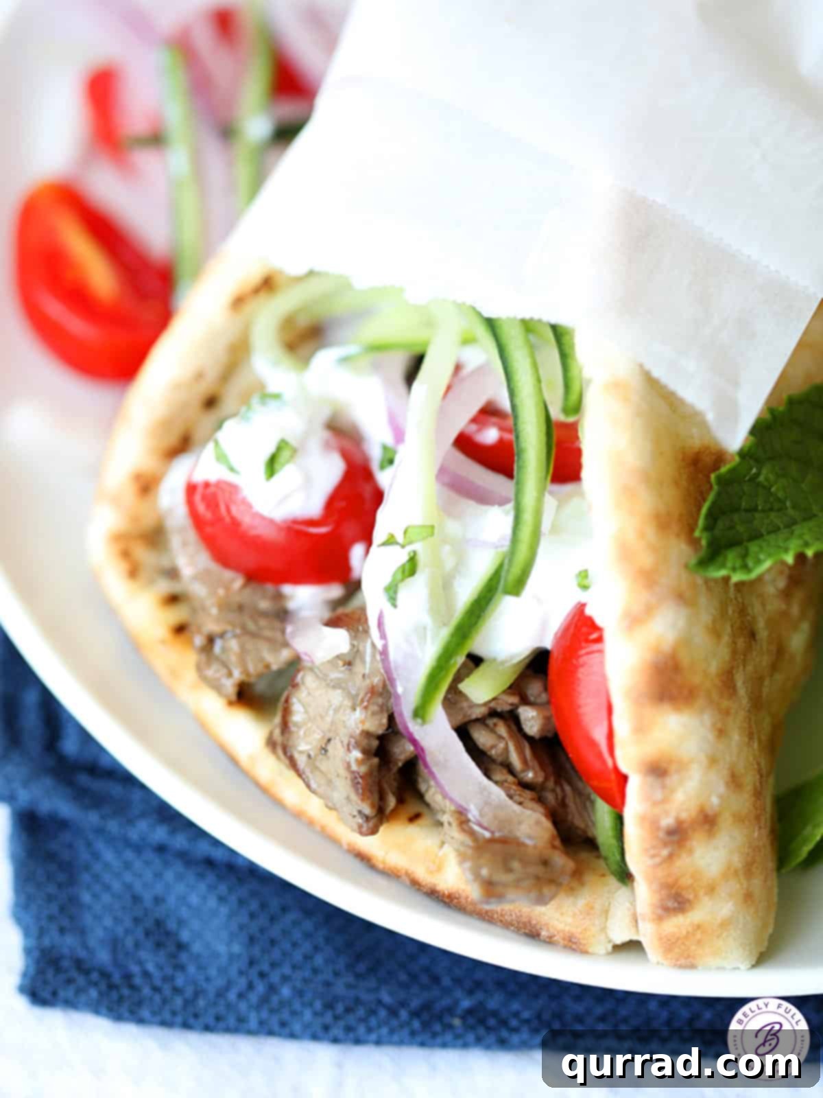 Close up view of assembled beef gyros, showing tender beef, fresh tomatoes, and cucumber in warm pita bread, topped with a generous drizzle of tzatziki sauce.