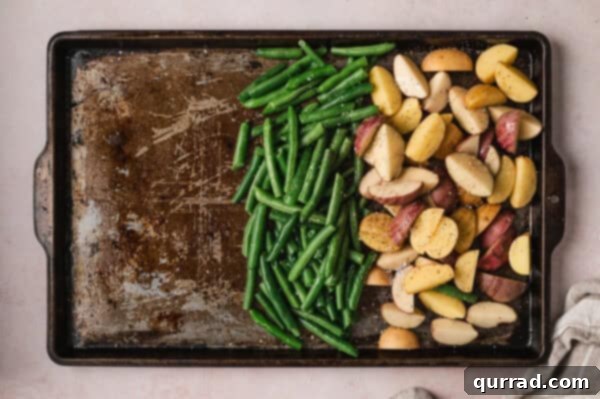 Fresh green beans and quartered potatoes on a baking sheet, seasoned with olive oil, salt, and pepper