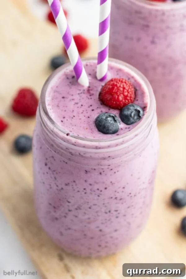 Vibrant blueberry smoothie in a tall glass, garnished with fresh blueberries, raspberries, and a stylish purple and white striped straw, set against a bright background.