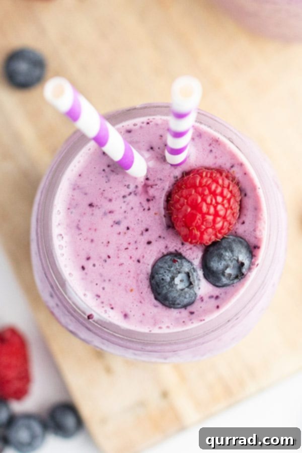 Overhead shot of a blueberry smoothie in a glass, garnished with two fresh blueberries and a single raspberry, highlighting its vibrant color and appealing presentation.
