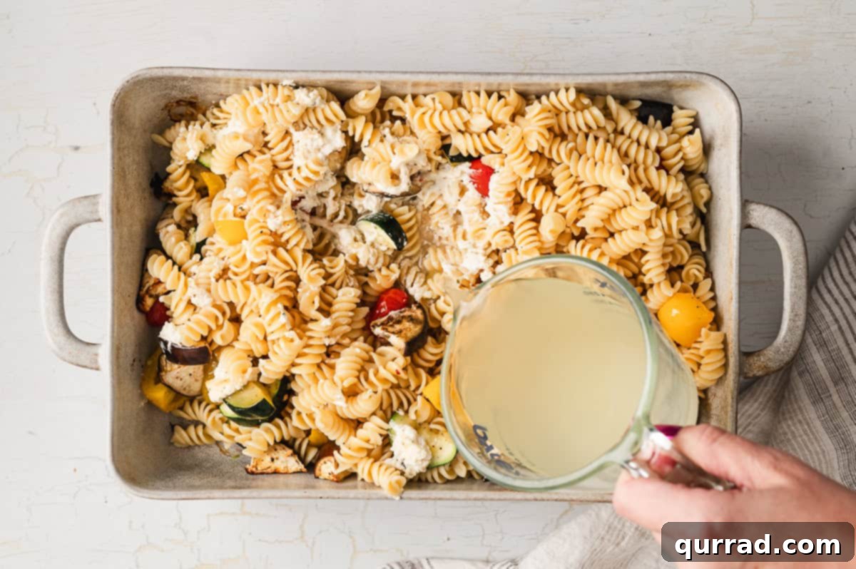 Velvety Ratatouille Pasta 7 A hand pouring pasta water over cooked rotini and roasted vegetables in a baking dish.