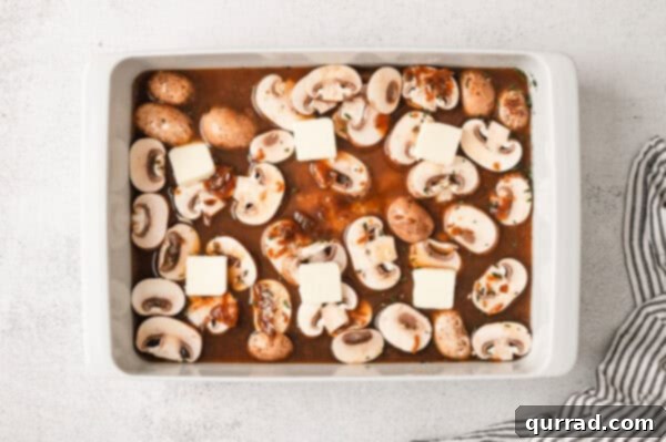 Sliced mushrooms in beef broth in a casserole dish with butter pats