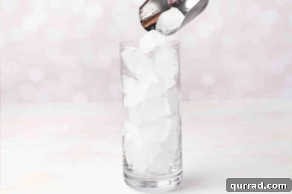 A hand pouring ice cubes into a tall highball glass, preparing for a cocktail.